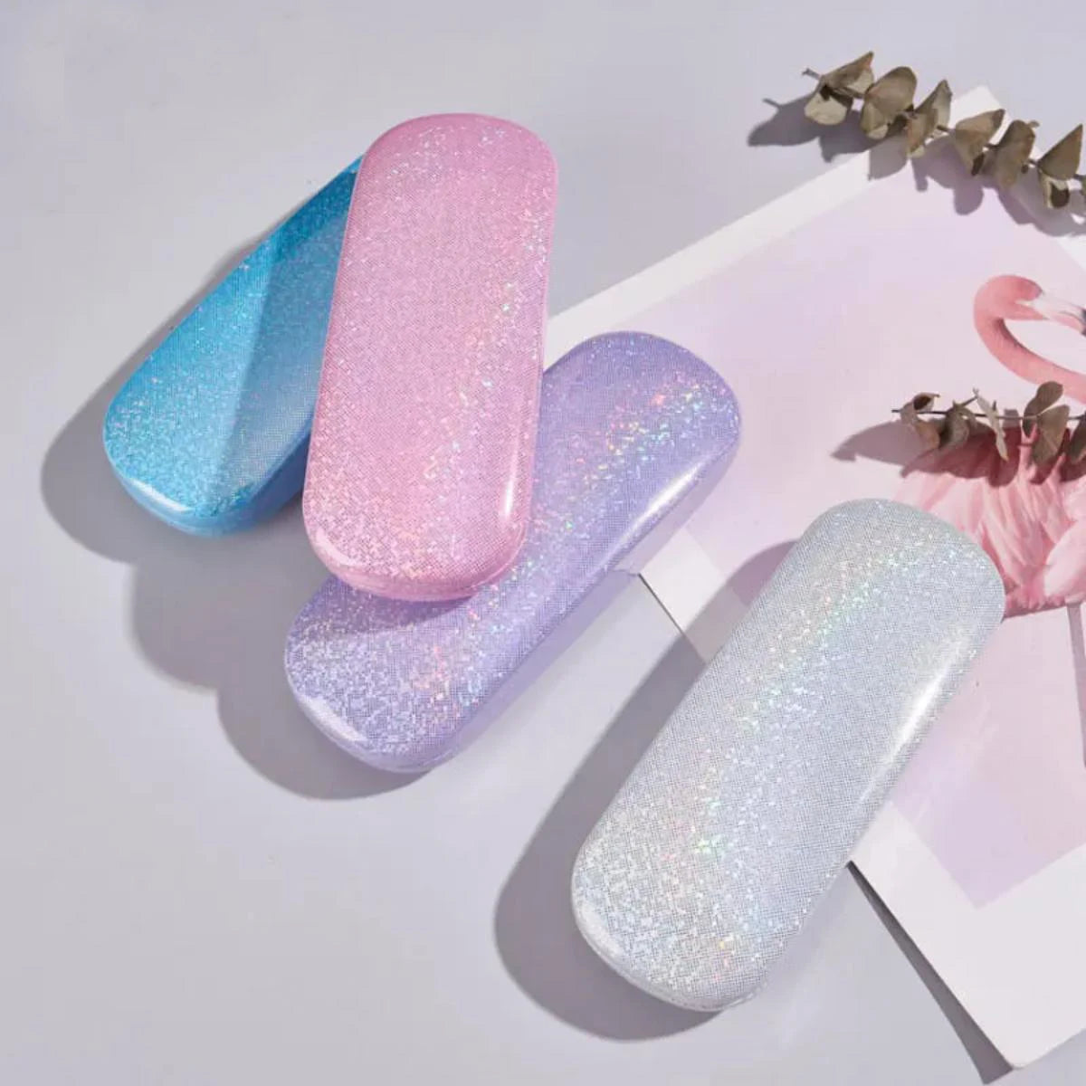 Une collection de cinq étuis pour lunettes à paillettes en bleu, rose et violet sont exposés sur un fond blanc