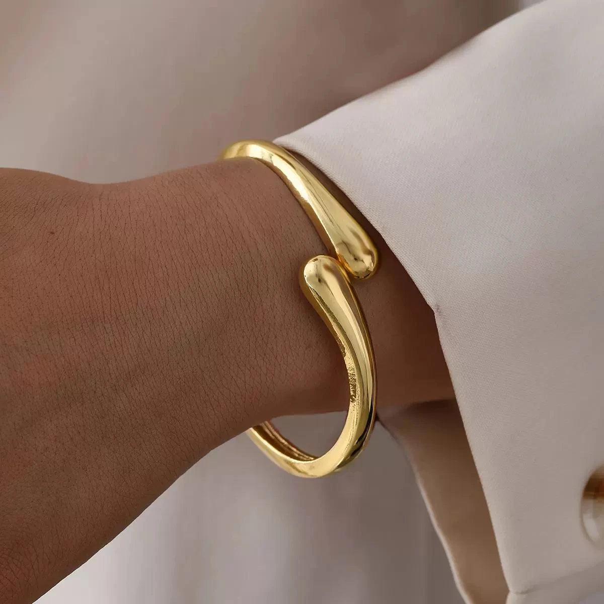 A close-up of a gold chunky cuff bangle bracelet worn on a wrist, next to a white sleeve.