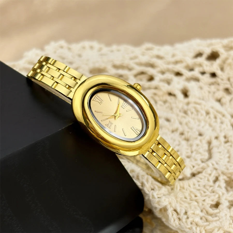 Reloj vintage dorado de esfera redonda y correa de metal sobre una superficie oscura junto a tela texturizada color crema.