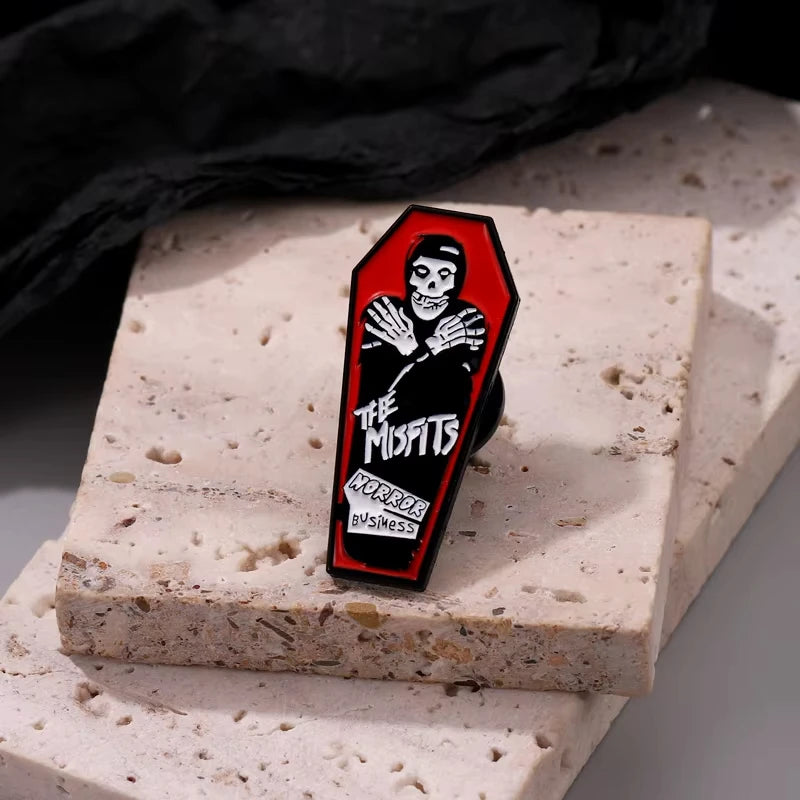 Schwarze Emaille-Anstecknadel mit Totenkopf-Design und dem Text The Misfits Horror Business auf einer hellrosa strukturierten