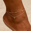 A gold geometric anklet bracelet with a heart charm worn on a person's ankle.