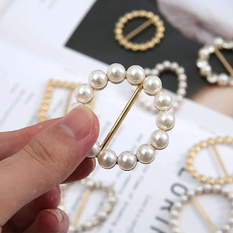 A hand holds a gold-toned button clip decorated with small white pearls and rhinestones.