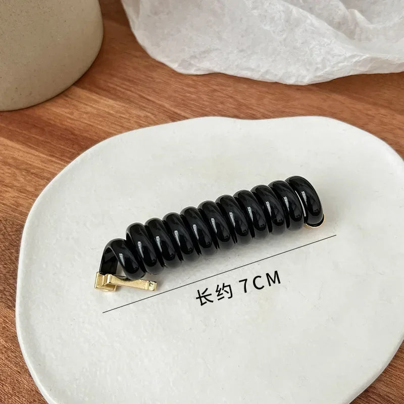 A black rubber braided ponytail holder is displayed on a white plate, with a measurement line indicating a length of 7cm.