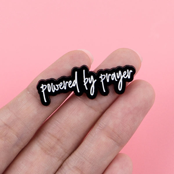 A hand holds a black enamel pin with the text powered by prayer on a pink background.