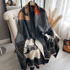 A luxury plaid cashmere feel scarf and shawl wrap in shades of black, gray, beige, and burnt orange draped over a white chair