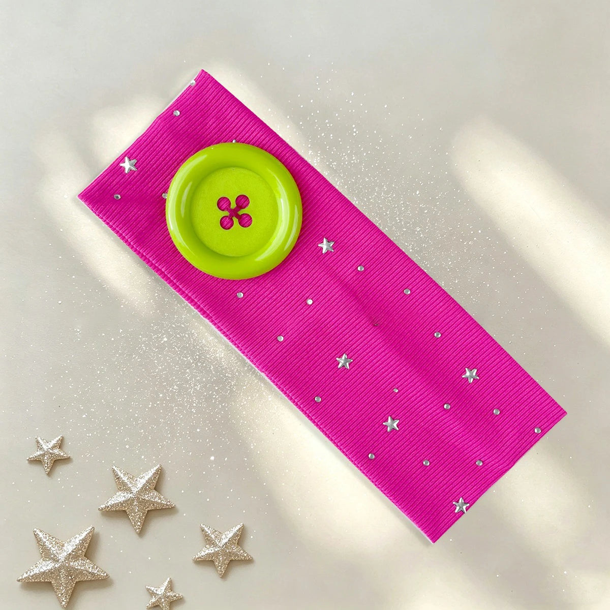 A bright pink headband with silver studs and a lime green button, lying on a light surface with scattered silver stars.