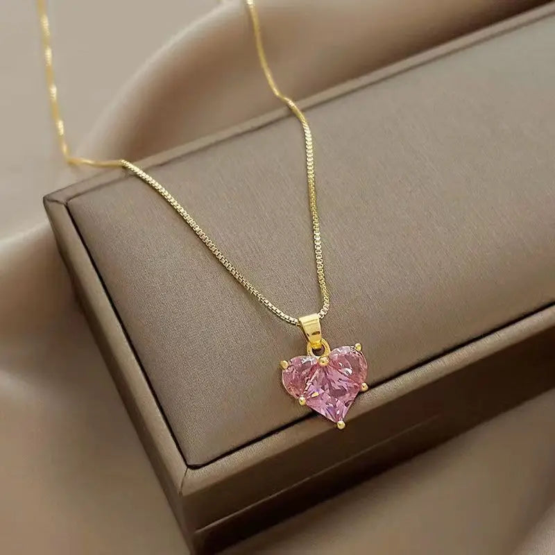 A delicate gold chain necklace featuring a pink crystal heart pendant displayed in a brown box.