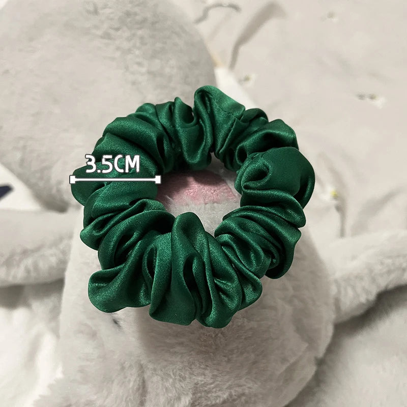 Goma para el pelo de tela verde esmeralda oscura hecha de seda fruncida, mostrada contra un fondo blanco esponjoso.