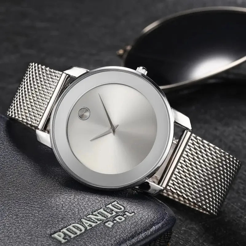 A silver quartz wristwatch with a mesh strap is shown resting on a dark surface next to a watch case.