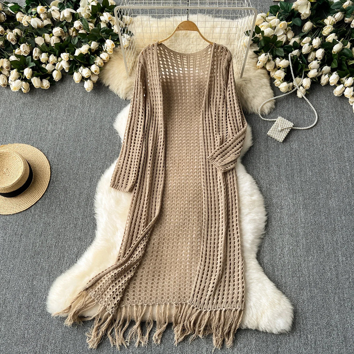 A beige knitted cardigan with fringe details draped over a white faux fur rug, surrounded by white flowers.