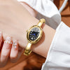 A close-up of a gold-toned quartz watch bracelet with a black face and blue hands worn on a wrist.