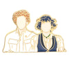 Broche émail dorée représentant deux figures stylisées aux cheveux bouclés et vêtements.