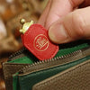 A hand is holding a red leather charm shaped like a turtle with a gold head, positioned near a green and brown leather wallet