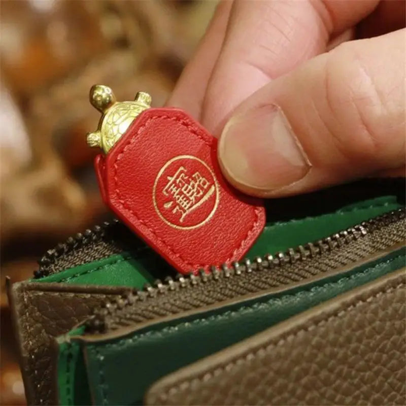 A hand is holding a red leather charm shaped like a turtle with a gold head, positioned near a green and brown leather wallet
