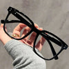 Una mano sostiene unas gafas oscuras cuadradas azules que bloquean la luz contra un fondo gris.