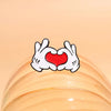 A white enamel pin depicting a cartoon hand gesture forming a heart shape with a red center.