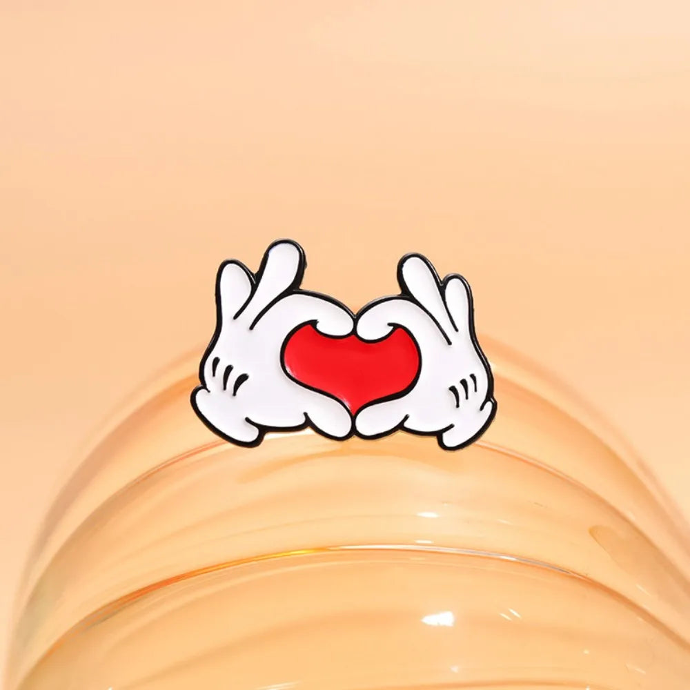 A white enamel pin depicting a cartoon hand gesture forming a heart shape with a red center.