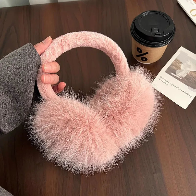 A pink faux fur earmuff is displayed on a wooden table next to a black cup and a piece of paper.