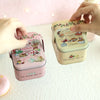 A hand is holding a pink decorative tinplate candy box with a carrying loop, featuring illustrations of treats and greenery.