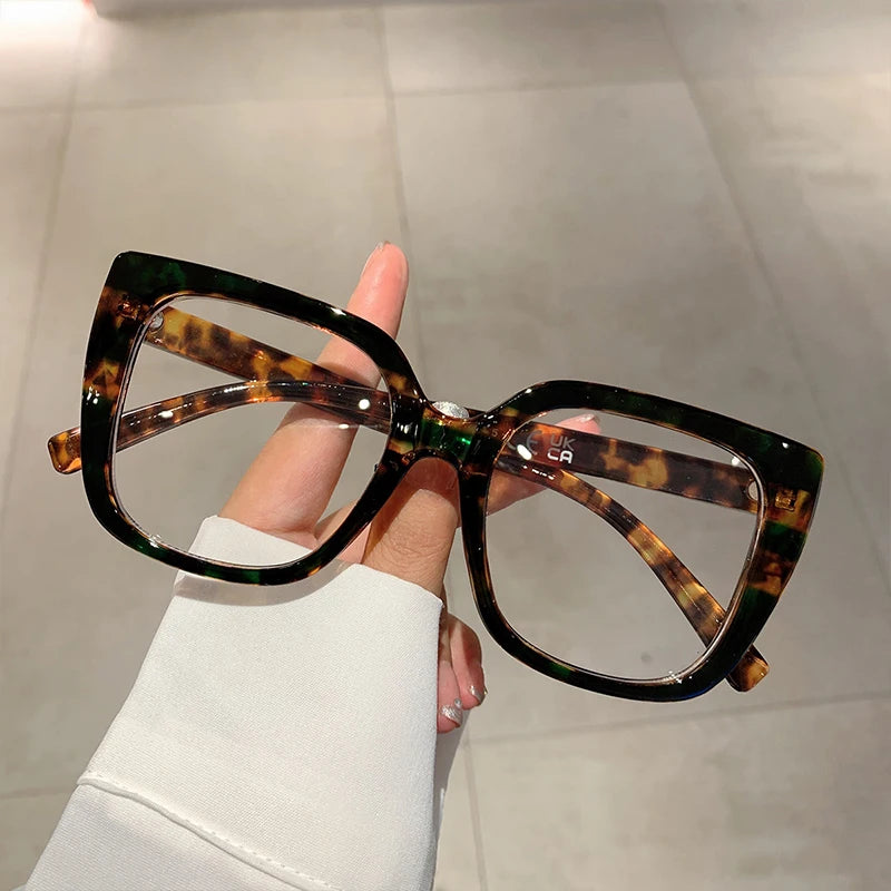 Eine Hand hält eine trendige, große, quadratische Brille mit braun-grüner Schildpattenfärbung.