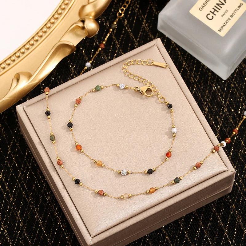 A gold-toned stainless steel beaded jewelry set with various colored beads displayed on a light brown box.