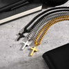 A collection of black, silver, and gold stainless steel Cuban link necklaces with cross pendants laid on a light gray surface