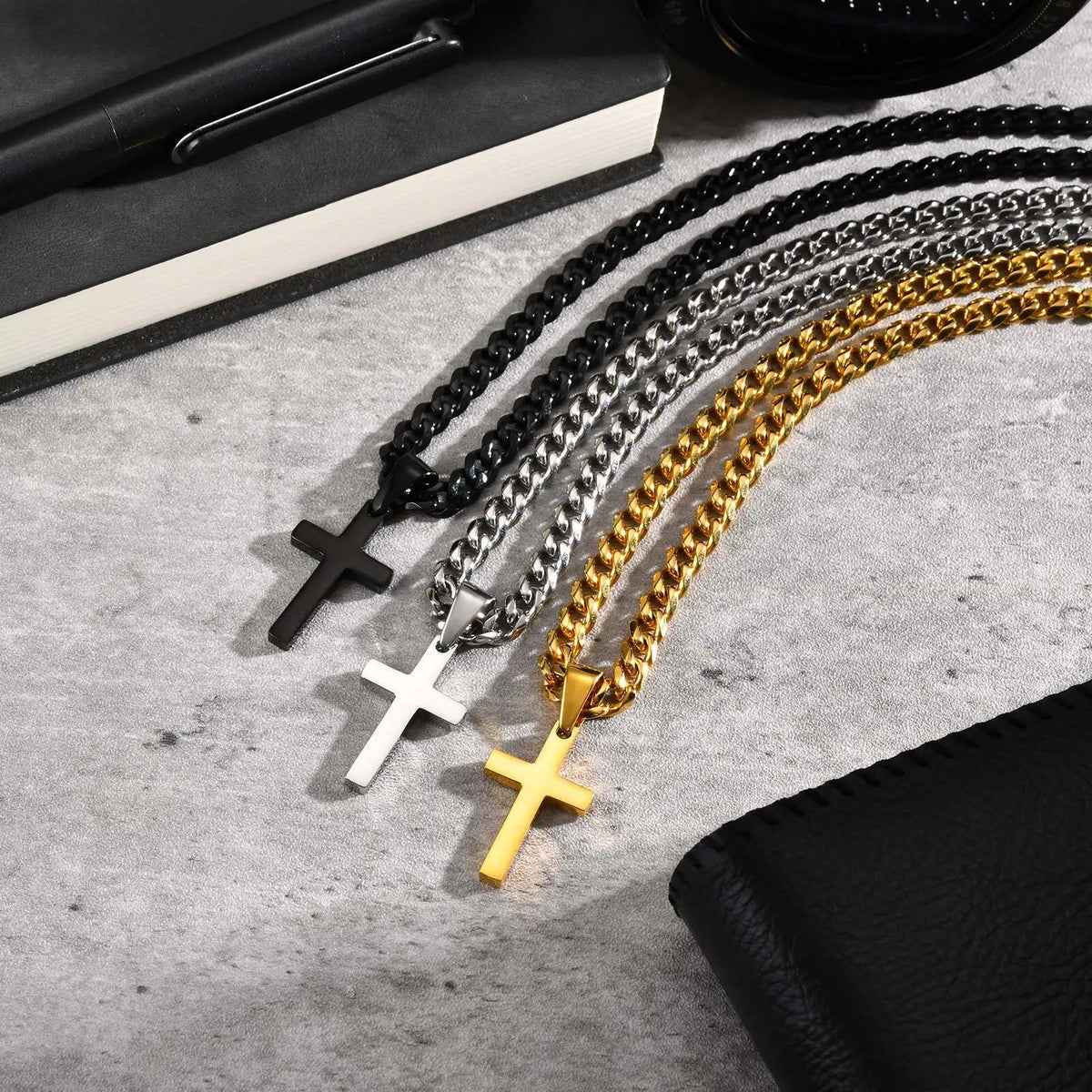 A collection of black, silver, and gold stainless steel Cuban link necklaces with cross pendants laid on a light gray surface