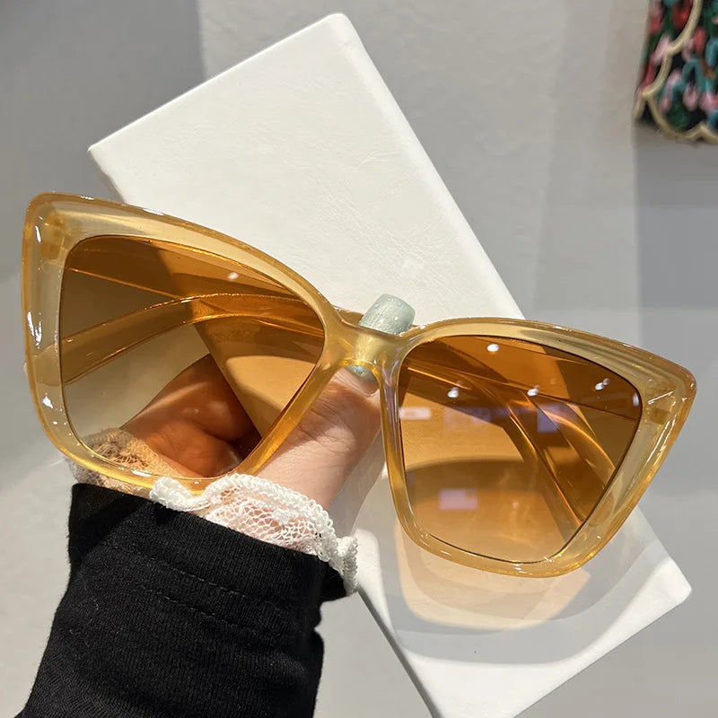 A pair of amber-tinted retro cat eye sunglasses being held by a hand against a white surface.