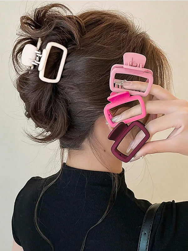 A hand is shown holding a set of square lightweight hair claw clips in various shades of pink and purple against dark hair.