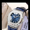 A close-up of a blue leather strap luxury watch with a silver case and blue dial on a light background.