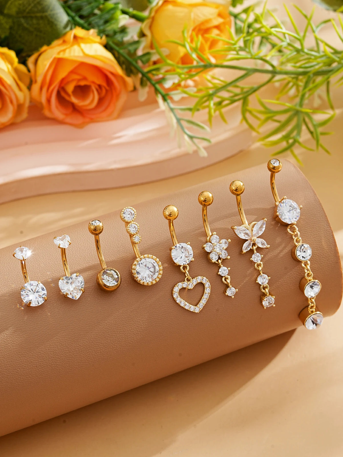 A set of gold dangle curved belly piercing jewelry featuring heart and flower designs displayed on a beige surface.