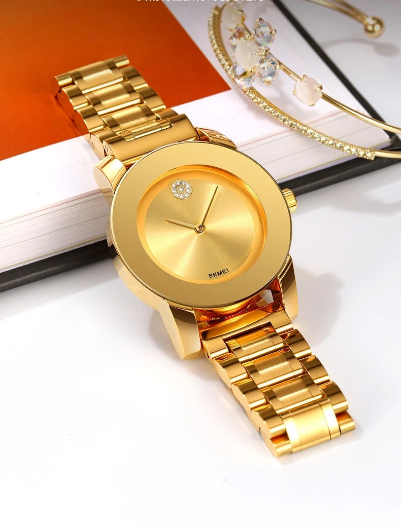 Reloj de cuarzo dorado con esfera redonda y joya brillante, sobre una superficie blanca.
