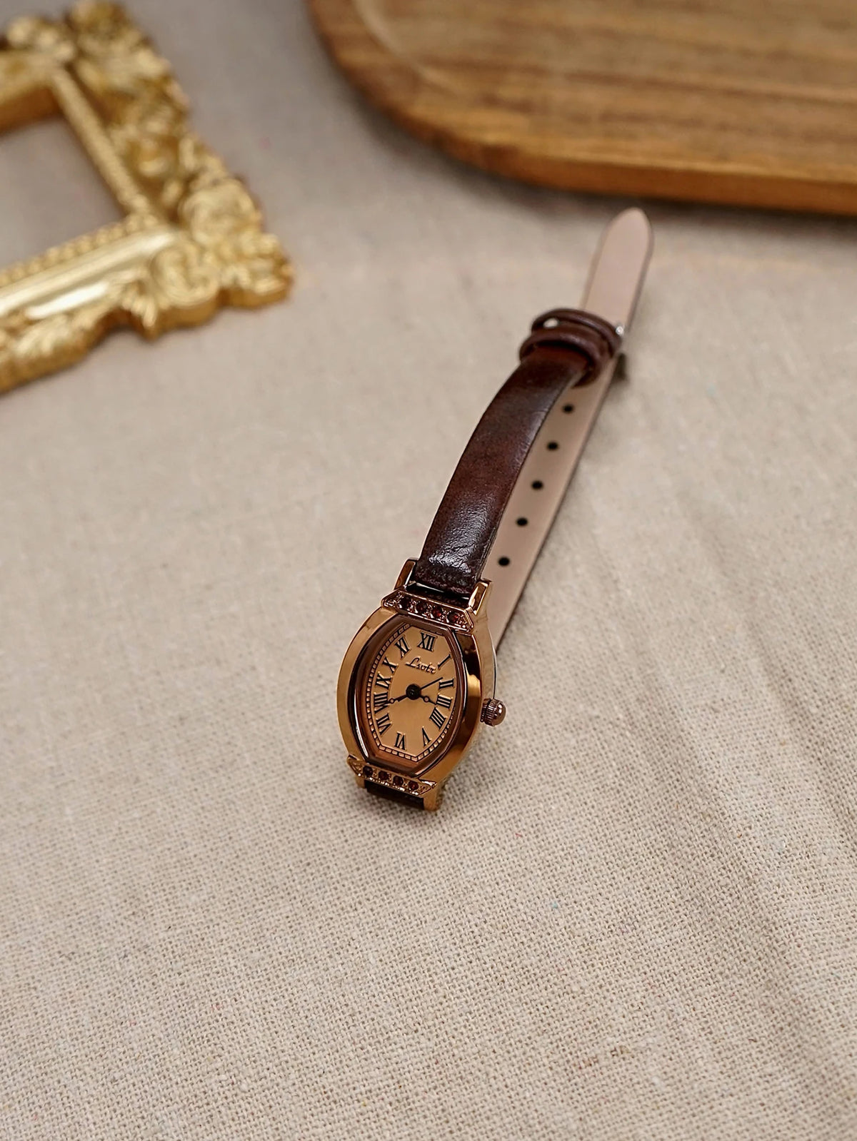Reloj de correa de cuero marrón retro con detalles dorados y pedrería sobre tela beige.