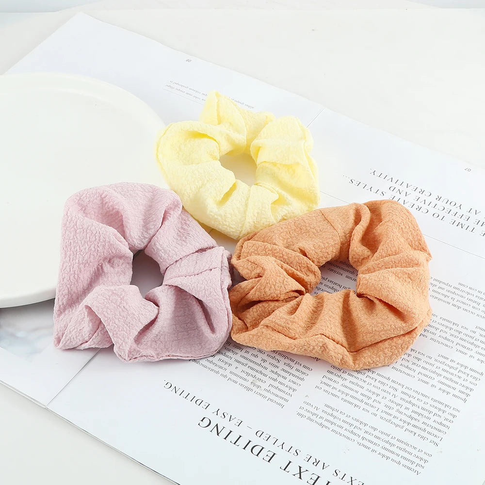 A set of three colorful knitted hair scrunchies in pink, yellow, and orange resting on a white paper.