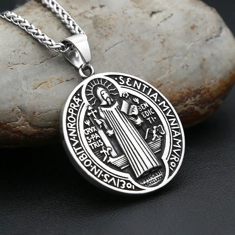A silver pendant necklace featuring an engraved Saint Benedict exorcism design, resting on a rough stone.