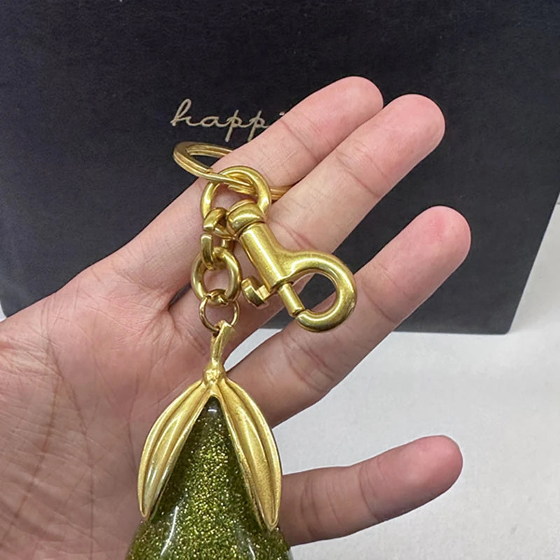 Un charm en forme de poire ton or avec une finition pailletée verte, tenu dans une main sur un fond sombre.