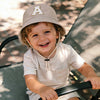 A smiling toddler wearing a beige letter print sun hat sitting on a dark outdoor chair.