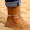A close-up of a woman's ankle showing an elegant geometric anklet bracelet on her skin against a sandy background.