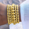 A set of gold round beaded bracelets stacked on a wrist against a light blue fabric background.