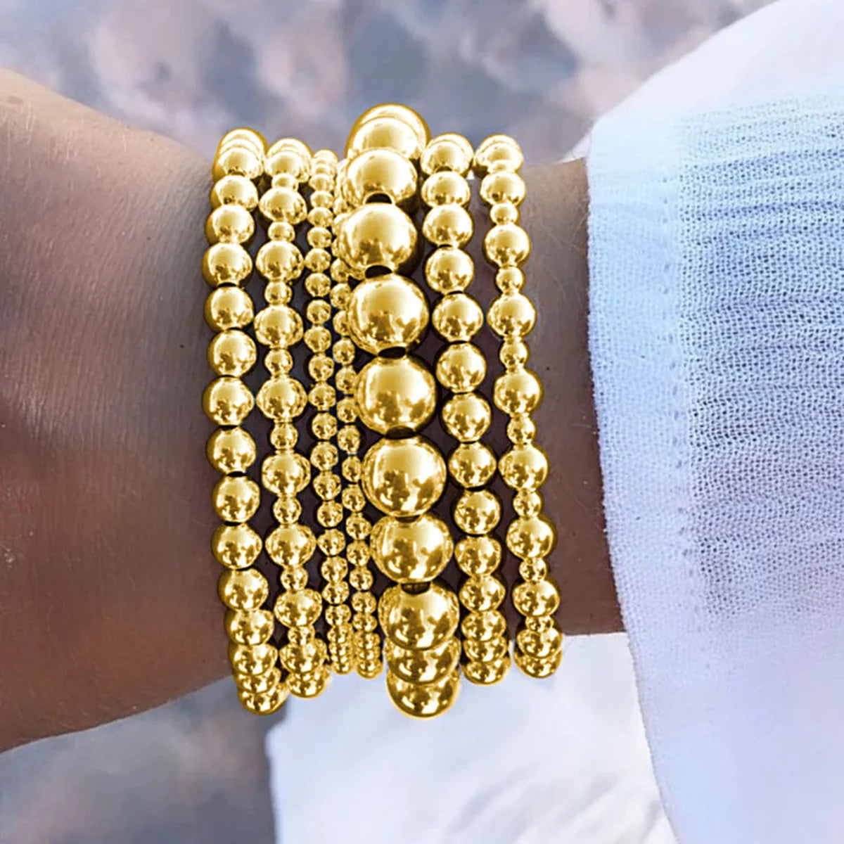 A set of gold round beaded bracelets stacked on a wrist against a light blue fabric background.