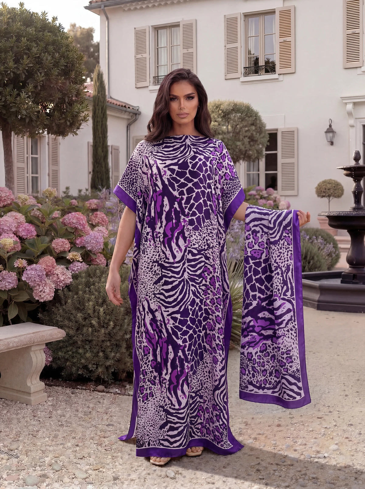 A woman wearing a purple and white leopard zebra print maxi kaftan standing outdoors in a garden setting.