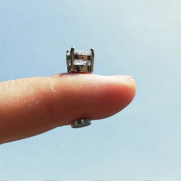 A hand holds a pair of silver magnetic stainless steel stud earrings against a light blue sky background.