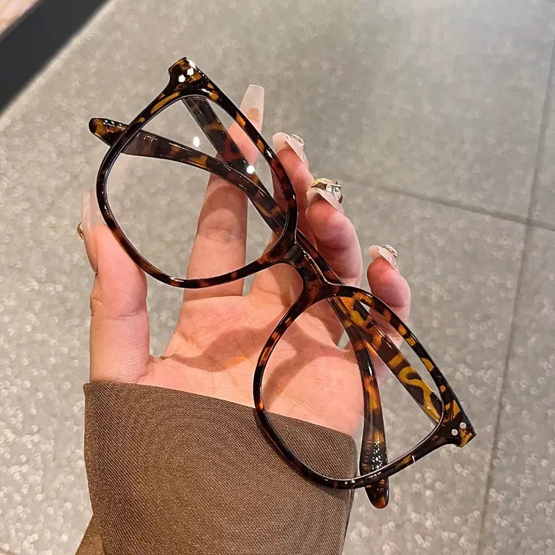 A hand holds a pair of tortoiseshell-patterned eyeglasses against a light gray background.