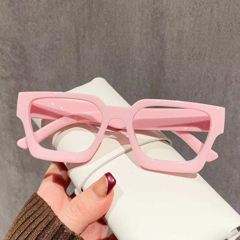 A hand holds a pair of pink square anti-blue light computer glasses against a white background.