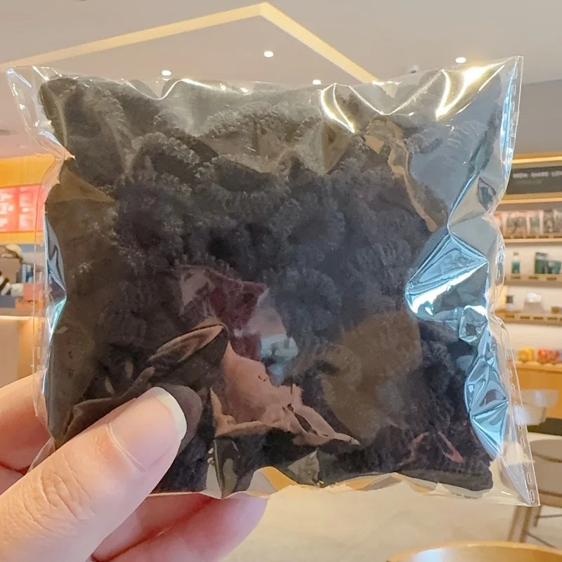 A hand holds a clear plastic bag filled with dark black elastic hair bands for girls.
