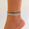 A stack of three beaded anklets in shades of beige, gold, and white worn on a person's ankle.