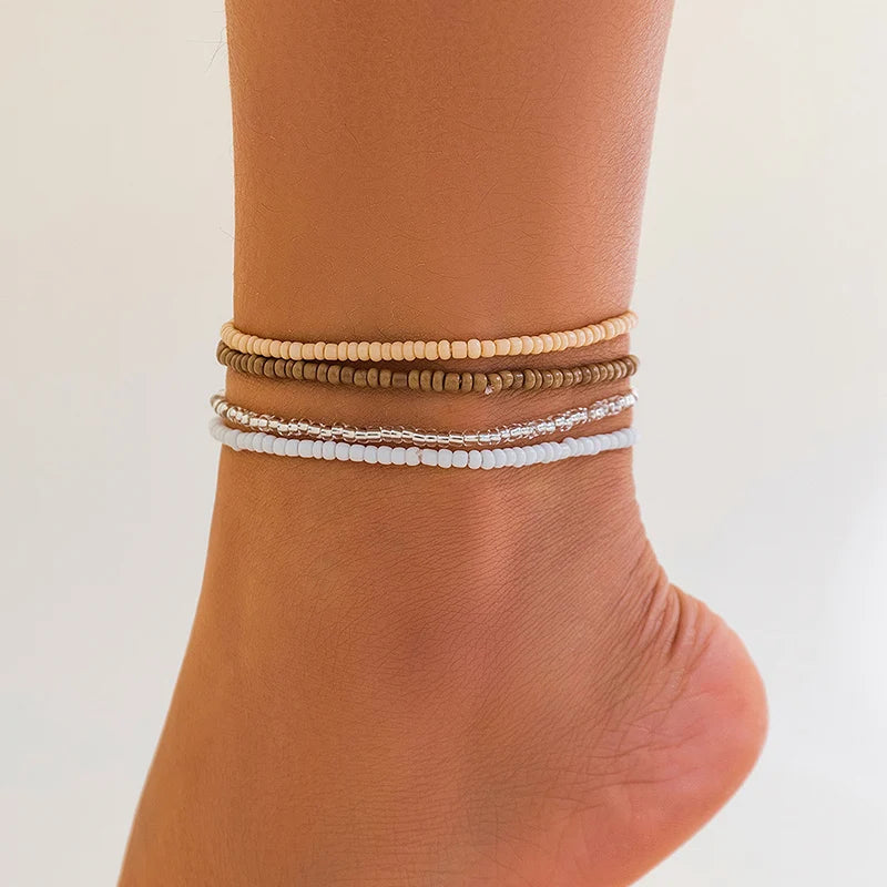 A stack of three beaded anklets in shades of beige, gold, and white worn on a person's ankle.