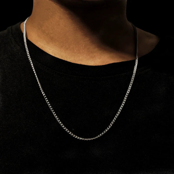 A close-up of a men's stainless steel Cuban link chain necklace worn around the neck against a dark background.