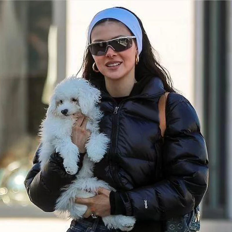 A woman wearing a black quilted jacket and sunglasses holds a white fluffy dog while wearing a blue knitted sports headband.