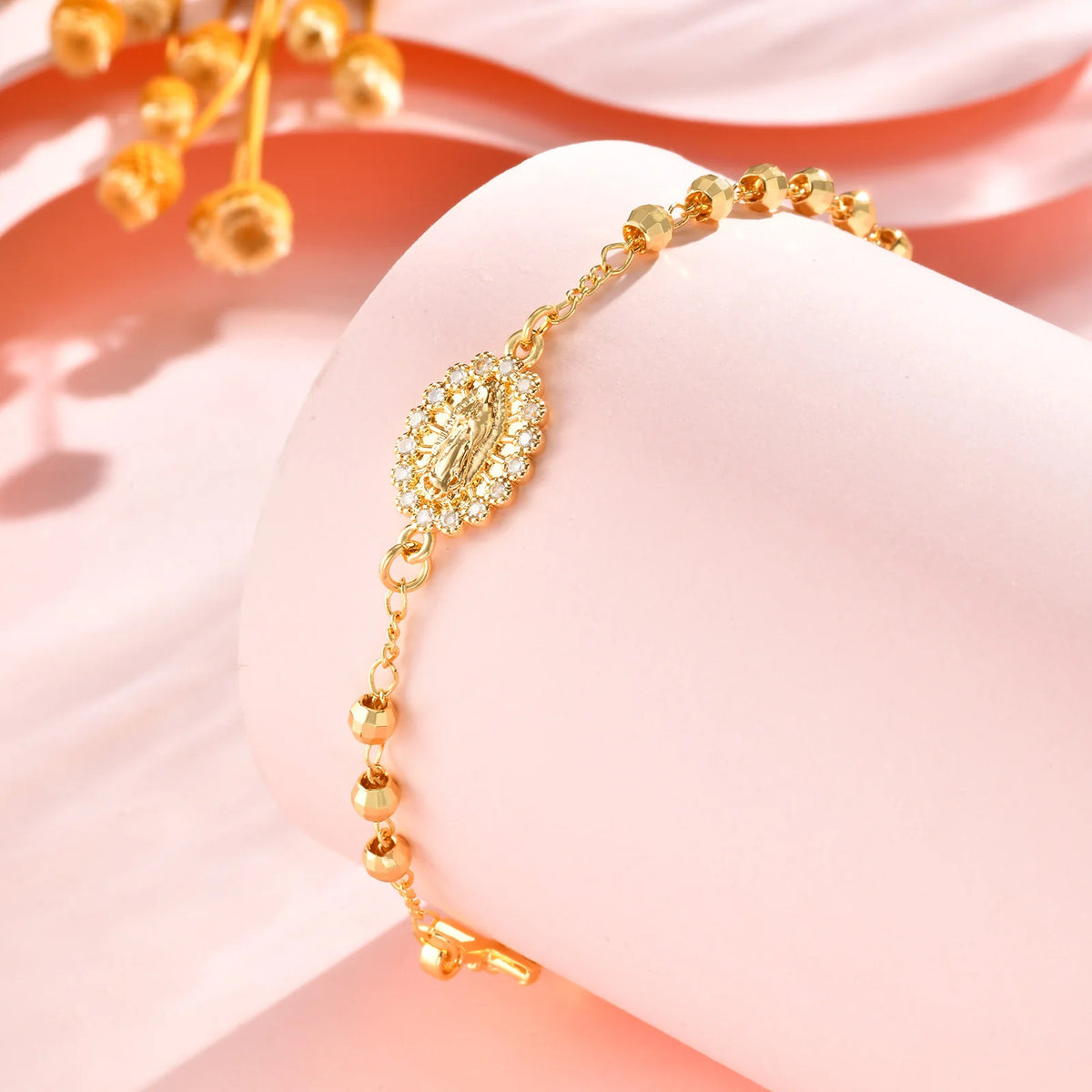 A close-up of an 18K gold plated rosary bracelet for women with a decorative pendant on a pink background.